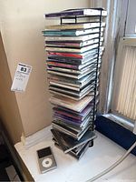 Vertical metal CD holder filled with over 30 music CDs, placed on a windowsill with an iPod classic lying next to it.