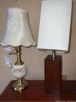 Photo showing both lamps side-by-side on wooden furniture: ceramic brass-tone lamp with floral fabric lampshade on left, wooden lamp with white rectangular lampshade on right.