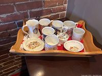Wooden tray holding multiple Royal Family commemorative items including mugs, plates, a trinket box, and a pitcher, showing overall lot contents and arrangement.