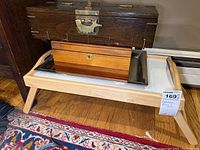 Large dark wooden box with metal latch placed behind two smaller wooden boxes on a silver metal tray which is set on wooden bed tray with handles and foldable legs.