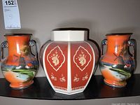 Front view of three ceramic vases together on a shelf, showing two matching orange Noritake vases with landscape scene and one hexagonal Lenox vase with red and floral panels.
