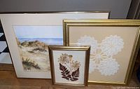 Three framed artworks visible: large signed watercolor with coastal landscape, smaller pressed flower collage, and medium-sized lace doily arrangement, all with gold frames.