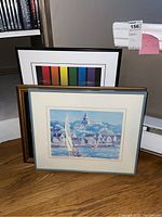 Three framed wall art pieces stacked on floor, Nantucket print with coastal homes and boats visible in front.