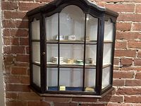 Front view of the wood and glass wall-mounted display case showing the arched top and three shelves with assorted small trinket boxes inside.