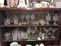Shelf display of multiple crystal glasses including tumblers, wine glasses, shot glasses, and martini glasses with detailed cut patterns, arranged on wood shelving.