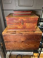 Stack of three wooden boxes: largest natural wood box on bottom, medium red painted box with latch in middle, and small red patterned decorative box on top.