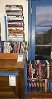 Stack of DVDs piled next to a wooden cabinet and blue door, showing many titles of movies and TV shows.