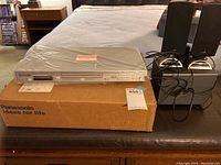 Panasonic DVD/CD player on top of its original box along with two black Insignia speakers on bases beside it.