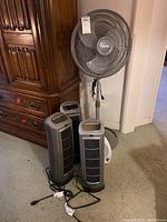 Photo of all five fans showing four Lasko tower fans and one Holmes pedestal fan arranged in a corner near wooden furniture.