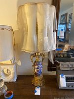 Front view of the vintage ornate painted lamp with fabric lampshade covered with plastic wrap, sitting on a wooden table.