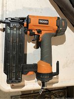 Front side view of Ridgid orange and gray brad nailer with metal nail magazine and air hose fitting attached.