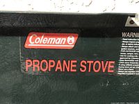 Close-up image of the Coleman logo and 'Propane Stove' printed on the stove's green hinged lid.