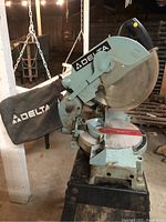 Side view of Delta chop saw showing dust collection bag and circular saw blade on pivot arm with handle, mounted on blue metal base.