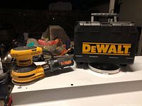 Yellow and black DeWalt orbital sander with dust collection bag and branded carrying case on table under low light.