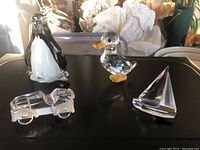Photo of four glass figurines on a black surface with some background clutter: penguin, duck, sailboat, car with missing tire.