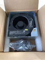 Top view of black ventilation fan unit inside original box with styrofoam packaging and plastic-wrapped component.