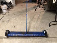 Wide front view of a blue magnetic floor sweep with a metal handle and two wheels on each end, positioned on a concrete floor.