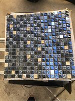 Square mosaic tile with reflective black, silver, and chrome small mirror pieces arranged in a decorative pattern.