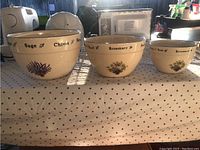 Three ceramic mixing bowls of different sizes placed side by side, decorated with herb names and botanical imagery on the exterior.