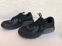 Pair of black Nike Air Max shoes angled view showing side and top of left and right shoes on white surface against textured off-white wall.