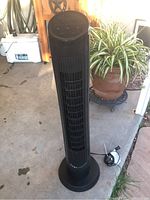 Full view of the black OmniBreeze oscillating tower fan standing on a concrete floor near a plant pot and cooler.