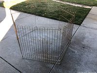 Photo showing metal pet exercise pen assembled on concrete with a gate panel latched closed. Six panels are connected to form a hexagonal shape.