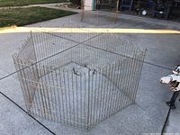 Hexagonal metal pet exercise pen assembled on a concrete driveway with 6 wire panels and curved tops visible, surrounding a central open area.