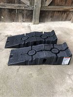 Two black plastic vehicle ramps placed side by side showing textured ribbed surface and circular reinforcements under natural light on concrete floor with wooden background.