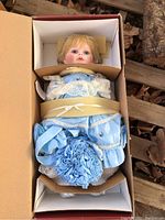 Full view of Sarah porcelain doll in box wearing blue dress with lace and floral accessory.