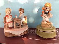 Photo shows two vintage music boxes placed side by side. The first depicts two children sitting at a table, the second a girl with a cat figurine on a green base.