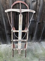 Front view of vintage wooden sled leaning against wooden fence. Visible curved handle and 'FLYING' brand on wood.