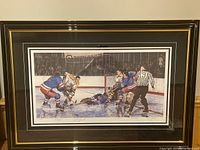 Framed hockey print featuring players in blue and yellow jerseys near the net, showing in-game ice action and a referee on the ice.