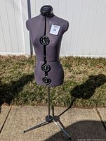 Full view of the adjustable dress form on its tripod metal stand outdoors on pavement near grass and white fence.