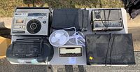 Photo showing seven electronic items on pavement including JVC CD boombox, Emerson stereo VCR, Toshiba multi-band radio cassette recorder, Realistic AM/FM radio, Pioneer CD player, GTE phone, and white clip-on fan.