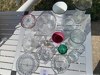 Top view of entire glassware and vase collection on white slatted table outdoors, showing variety of shapes and sizes including clear and colored glass items.