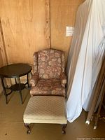 Floral wing back chair with matching ottoman and adjacent dark wood round scallop occasional table in a storage setting.