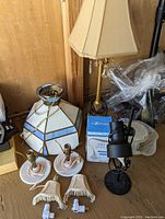 Photo showing tall candlestick table lamp with beige shade and crystals, blue and white glass hanging lamp shade, black desk lamp, white plates under plastic, two nightlight shades, and electric adapters on wooden floor.