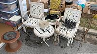 Two white metal outdoor glider chairs with decorative cutout backs and seats, visible rust. Round white metal outdoor table and other metal garden accessories arranged around the chairs.
