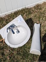 White pedestal sink basin and separate pedestal base placed on grass outdoors. Sink has chrome faucet and appears lightly used.