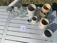 Six beer steins and mugs displayed on a white outdoor table, including clear glass steins with metal lids and various decorated stoneware steins.