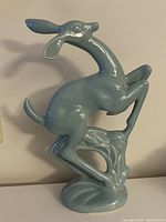 Front and side views of light blue ceramic fawn deer figurine with elongated ears and legs in a playful stance on a molded base.