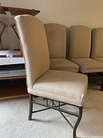 Photo showing side and front angle of multiple beige upholstered dining chairs with metal bases, arranged in a row inside a room.