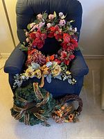 Large wreath with red poinsettias, purple flowers, green leaves, and bow displayed on blue chair alongside other wreaths.