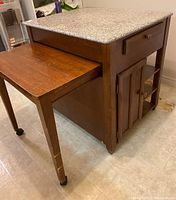 Front and side angle of kitchen island showing marble top, extended wooden table with wheels, drawer, cabinet, and shelving.