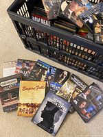 Photo showing the black plastic storage bin mostly filled with DVDs and several DVDs displayed outside the bin on the carpet, showing titles such as Empire Falls, The Girl with the Dragon Tattoo, Die Hard, and others.