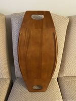 Top view of surfboard-shaped wooden tray resting on a chair, showing wood grain, finish, and cutout handles.
