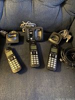 Photo of three Panasonic cordless handsets, three base units with docking stations, charging cables and power adapters on a dark fabric surface.
