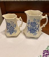 Pair of two ceramic pitchers placed on a white cloth-topped surface, showing blue floral designs on cream background and distinctive hexagonal shapes with sculpted handles.