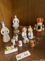 Full view of all figurines on a wooden shelf, including traditional female porcelain figurines, white horses, flowers, glass vessel, Red Cross thimble, mouse and wooden nutcracker.