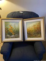 Pair of framed floral paintings leaning against a chair, showing full front view of each painting and frame.